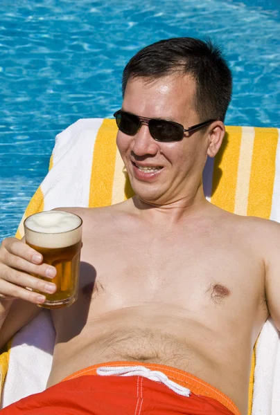 Homem segurando uma cerveja em uma espreguiçadeira ao lado de uma piscina — Fotografia de Stock