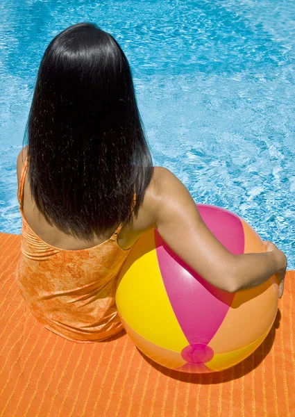 Mujer con el pelo largo y negro sentada junto a la piscina sosteniendo una pelota de playa — Foto de Stock