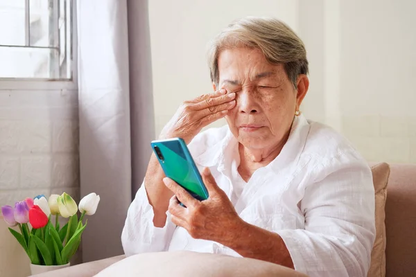 白髪のアジア系の高齢女性は携帯電話を使い 手で目をこすっている 高齢者の目の問題 ぼやけたビジョン ストック画像