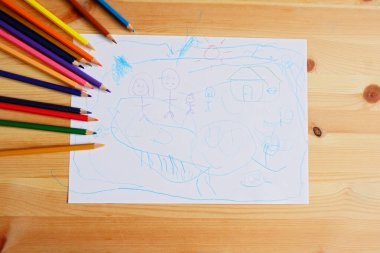 Mutlu bir aile ve ev çizen sevimli Asyalı çocuk. Ahşap masa üzerinde renkli kalemler ve beyaz kağıt ile, üst görünüm