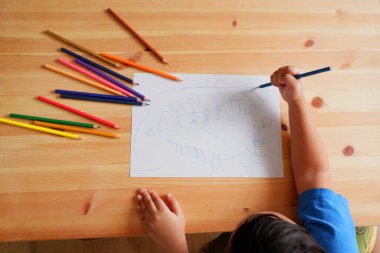 Mutlu bir aile ve ev çizen sevimli Asyalı çocuk. Ahşap masa üzerinde renkli kalemler ve beyaz kağıt ile, üst görünüm