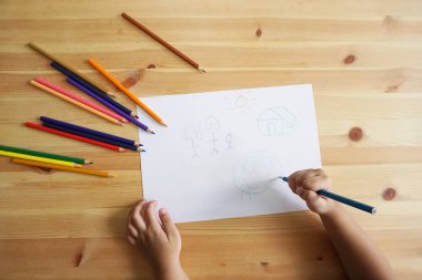 Mutlu bir aile ve ev çizen sevimli Asyalı çocuk. Ahşap masa üzerinde renkli kalemler ve beyaz kağıt ile, üst görünüm
