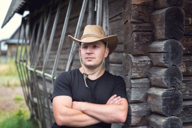 Ahşap bir evin arka planında şapkalı ve siyah tişörtlü bir kovboy..