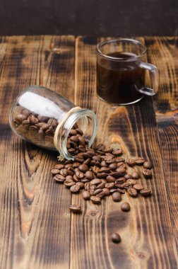 Bir fincan kahve ve fasulye, ahşap bir masanın üzerindeki cam kavanozdan serpilmiştir..