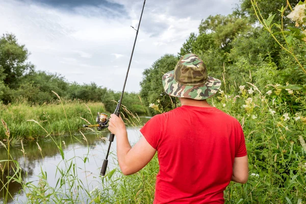 Panama şapkalı bir balıkçı ve kırmızı tişörtlü bir olta kamışı kullanıyor. Arkadan bak. Hobi balıkçılık.