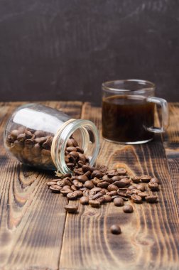 Bir fincan kahve ve fasulye, ahşap bir masanın üzerindeki cam kavanozdan serpilmiştir..