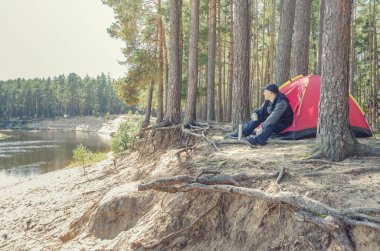 Genç adam kayadaki ormanda bir çadırın yanında oturuyor. Kamp..