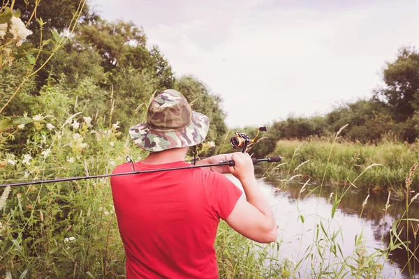 Fisherman in a panama hat and a red t-shirt casts a spinning rod. View from the back. Hobby fishing.