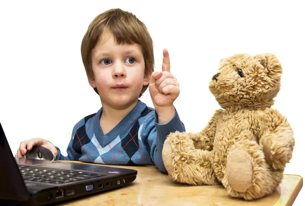 O menino ensina um urso de brinquedo na frente do laptop — Fotografia de Stock