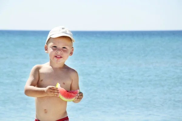 Frumos băiat care mănâncă felie de pepene verde pe plajă — Fotografie, imagine de stoc