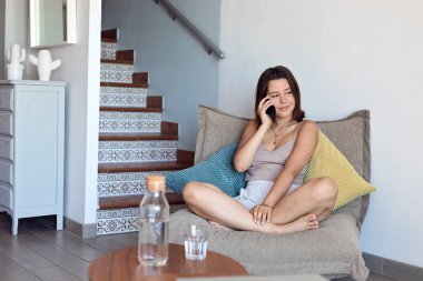 Yazlık kıyafetli genç kız evde telefonla konuşuyor. Rahat ve rahat bir kız zamanı. Gülümsüyor, evin içinde rahatlatıcı atmosferin tadını çıkarıyor. 