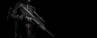 Siyah elbiseli bir adam elinde otomatik bir karabina tutuyor. Yüzü maskeyle kaplı, elleri eldivenli. Karanlık bir arka planda silahı olan bir figür..