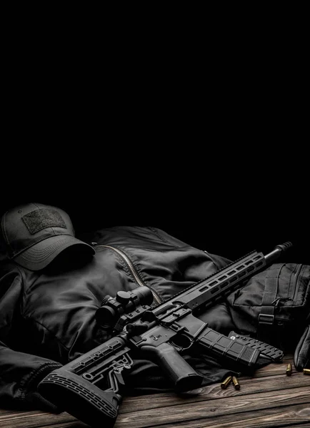 Modern automatic rifle with a telescopic sight on a dark background. Jacket, cap, gloves on a wooden table. The uniform of a guard or a mercenary.
