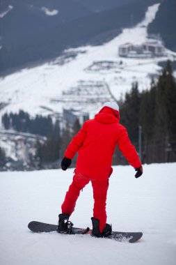 Kırmızı ceketli, snowboard 'lu bir adam bir dağ yamacında duruyor..