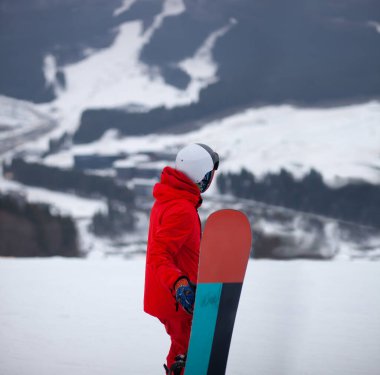 Kırmızı ceketli, snowboard 'lu bir adam bir dağ yamacında duruyor..
