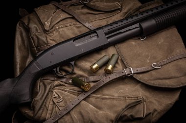 Siyah bir 12 kalibrelik pompalı tüfek ve cephane sırt çantasında. Plastik malzemeli yumuşak delikli bir silah. Koyu arkaplan.