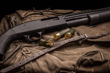 Siyah bir 12 kalibrelik pompalı tüfek ve cephane sırt çantasında. Plastik malzemeli yumuşak delikli bir silah. Koyu arkaplan.