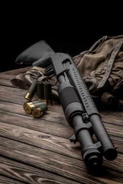 Siyah bir 12 kalibrelik pompalı tüfek ve cephane sırt çantasında. Plastik malzemeli yumuşak delikli bir silah. Koyu arkaplan.