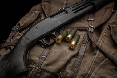 Siyah bir 12 kalibrelik pompalı tüfek ve cephane sırt çantasında. Plastik malzemeli yumuşak delikli bir silah. Koyu arkaplan.