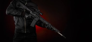 Siyah elbiseli bir adam elinde otomatik bir karabina tutuyor. Yüzü maskeyle kaplı, elleri eldivenli. Karanlık bir arkaplanda kırmızı ışıklı bir silah taşıyan bir figür..