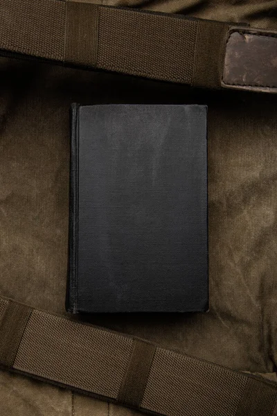 A closed book with a black cover on an old hiking backpack. Vintage khaki canvas background.