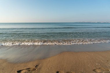 Açık güneşli bir günde kumlu bir sahilde dalgalar akıyor. Arka planda şehir.