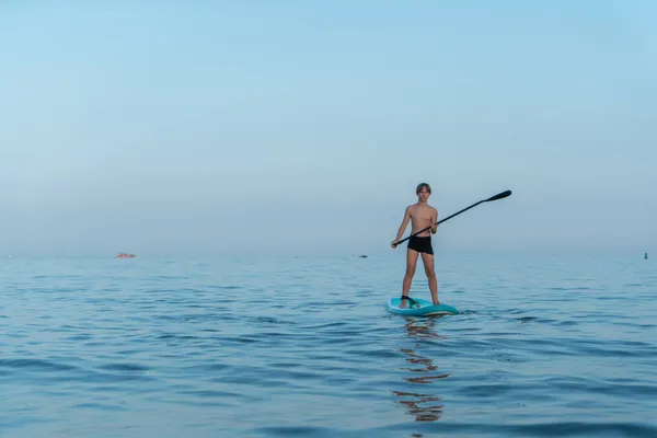Een jongen van 11 jaar oud zwemt op een SUP board in de zee na zonsondergang. — Stockfoto