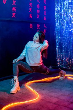 Şık bir genç kızın portresi, kapüşonlu bir kapüşonluyla dans eden ve saçlarını uzatan, neon bir arka plan. Spor, sigara, esneme, antrenör, egzersiz, sicim.