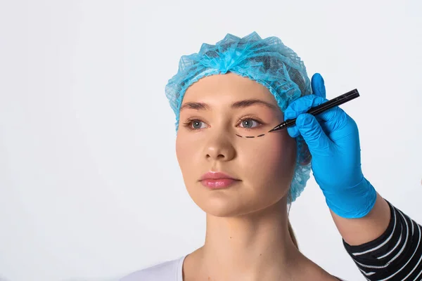 与医生的手戴在脸上 整形外科概念画穿孔线蓝色手套在工作室的背景下 戴蓝色医疗帽子的黑眉毛的漂亮女孩 — 图库照片