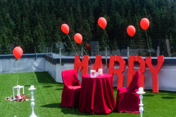 Red chairs and a tablecloth against the background of capital letters will you marry me. Red balloons and forest on the background