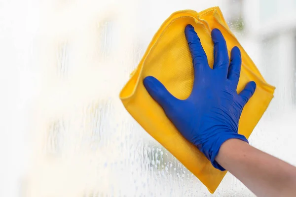 A woman or man washes a window at home. House cleaning. Washing dirty window glass detergent for window washing wearing blue mittens and a yellow cloth rag
