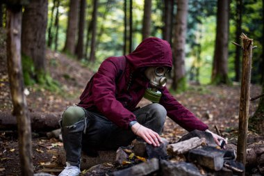 a male in a gas mask and cherry jacket sits in the middle of the forest. nuclear war concept
