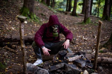 a man in a gas mask and cherry jacket sits in the middle of the forest. nuclear war concept