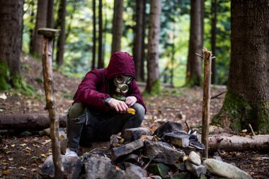 a guy in a gas mask and a cherry jacket sits in the middle of the forest and starts a fire. nuclear war concept