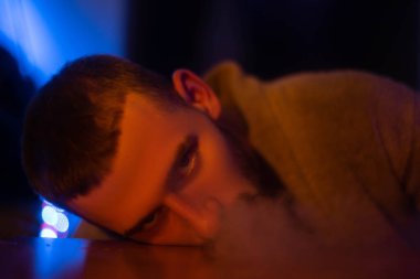 Şık bir genç adam kafasını masaya koyar ve kameraya vahşice bakar. Gri duman solur. Kapat..