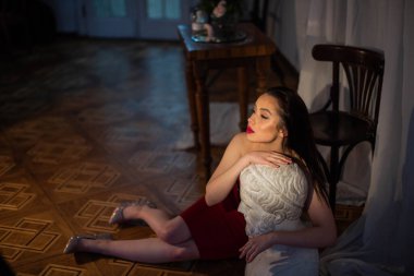 Kırmızı gece elbisesi ve parlak ayakkabıları olan muhteşem bir esmer manken. Güzel, çekici, seksi bir kadın kameranın önünde güzel bacaklarıyla heykellerin yanında oturuyor. Heykele sarılmış yan tarafa bakıyor..