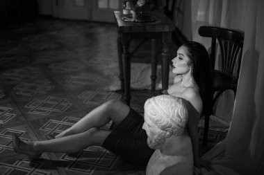 Kırmızı gece elbisesi ve parlak ayakkabıları olan muhteşem bir esmer manken. Güzel, çekici, seksi bir kadın kameranın önünde güzel bacaklarıyla heykellerin yanında oturuyor. Önüne bak. Gri bukle.