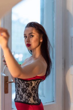 Uzun saçlı ve kıvrımlı şekilli genç bir Kafkasyalı. Bir bardak şarapla pencerenin yanında duruyor. Pencerenin arka planındaki kameraya bak..