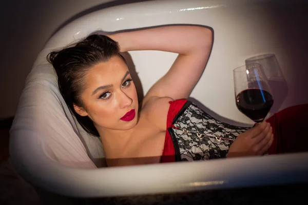 一个褐色眼睛的黑发女人 有着明亮的妆容 穿着红色连衣裙和紧身衣 躺在浴缸里喝着一杯红酒 喝了一浴酒 非常残忍 — 图库照片