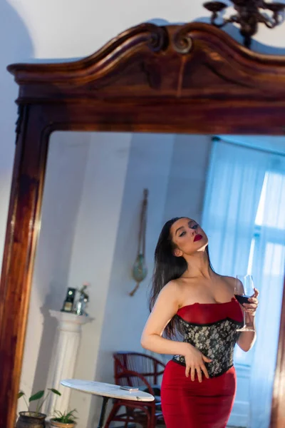 豪華な姿の赤いドレスを着た女性が鏡に映る姿を賞賛している 白い壁と家具のあるリビングルームの背景に立つ — ストック写真