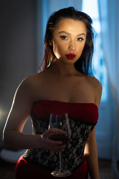 一个漂亮的姑娘 棕色的眼睛 黑头发 穿着红色的连衣裙 手上拿着一杯葡萄酒 在一个白色窗户的后面 看着摄像机 — 图库照片