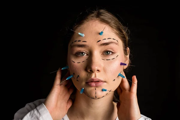 young beautiful girl with deep eyes looking into the camera. needles from syringes and arrows on her face. the girl touches her head with her hands. imagination of plastic surgery. on a black background.