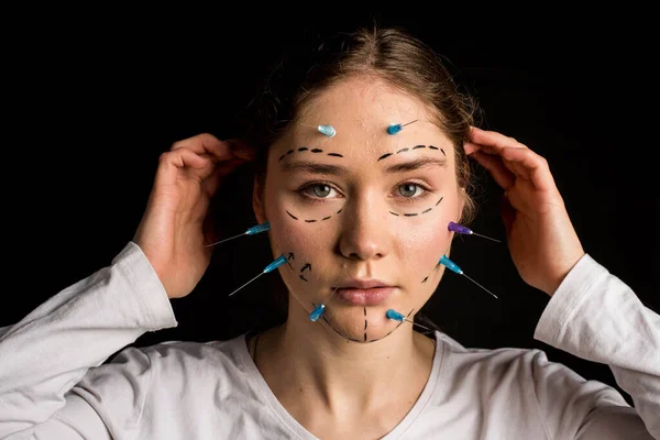 young beautiful girl with deep eyes looking into the camera. needles from syringes and arrows on her face. imagination of plastic surgery. on a black background.