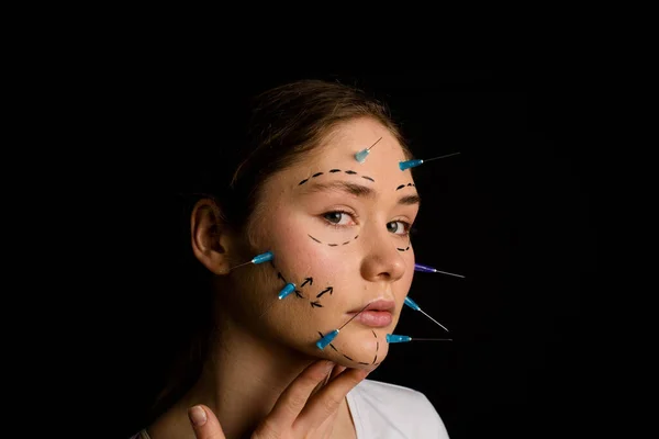 young beautiful girl with deep eyes looking into the camera. needles from syringes and arrows are drawn with a marker on her face. imagination of plastic surgery. the girl touches her neck. on a black background.