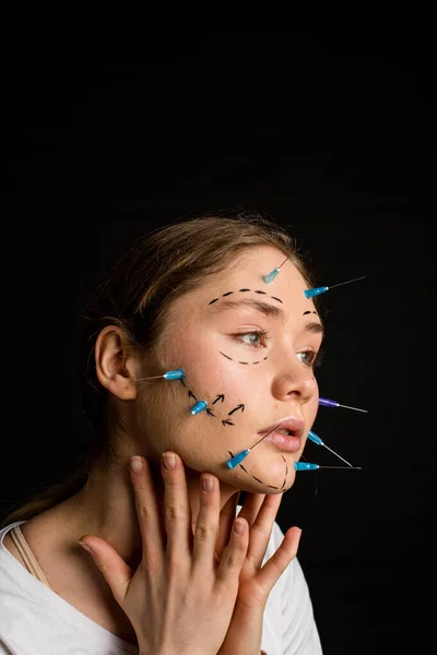 young beautiful girl with deep eyes looking into the camera. needles from syringes and arrows are drawn with a marker on her face. the girl touches her neck. on a black background.