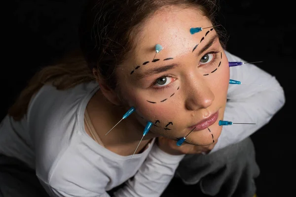 young beautiful girl with deep eyes looking into the camera. needles from syringes and arrows are drawn with a marker on her face. imagination of plastic surgery. the girl sits on the ground and looks into the camera. on a black background. close up