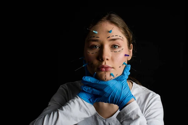 young beautiful girl with beautiful eyes looking into the camera. needles from syringes and arrows are drawn with a marker on her face. the girl touches her face with hands in gloves. on a black background.