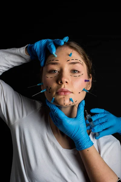 young beautiful girl with deep eyes looking into the camera. needles from syringes and arrows are drawn with a marker on her face. the girl touches her face with hands in blue gloves. on a black background.