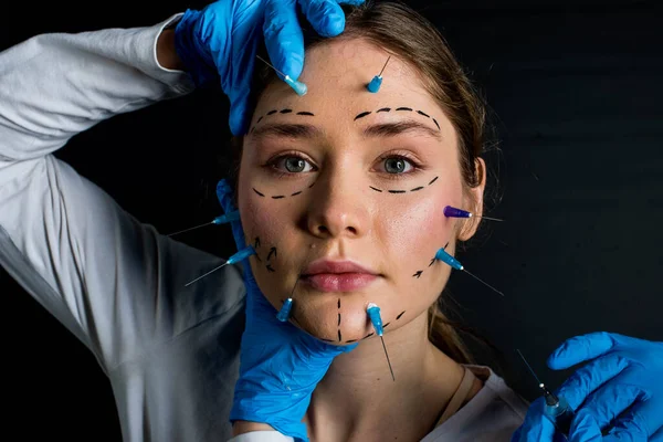 young beautiful girl with deep eyes looking into the camera. needles from syringes and arrows are drawn with a marker on her face. the girl touches her face with hands in blue gloves. on a black background. Close up