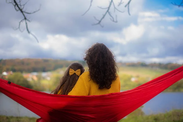 头发卷曲的母亲和头发上有蝴蝶结的女儿 都穿着黄色的毛衣坐在蓝色的吊床上 看着河水 — 图库照片